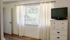 Een kamer met een raam, gordijnen, een televisie op een dressoir en houten vloeren.