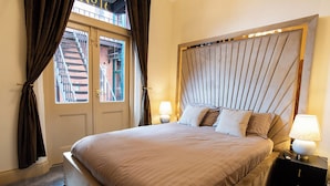 Habitación de hotel con una cama grande, una mesita de noche con lámpara, una ventana con cortinas y vistas a un edificio.