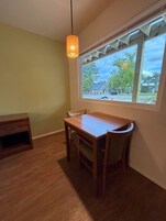 Un comedor con una mesa y sillas de madera, una lámpara colgante y una ventana con vista a una casa y árboles.