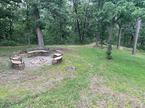 Un espacio al aire libre rodeado de bosque, con un brasero, bancos y un pequeño claro.