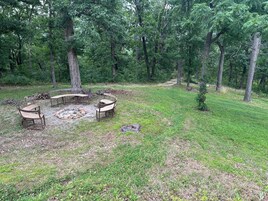 Un espacio al aire libre rodeado de bosque, con un brasero, bancos y un pequeño claro.