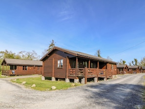 A rustic log cabin with a porch, a gravel driveway, and a clear blue sky. - Lodge 7 - Bron Eifion (Criccieth)