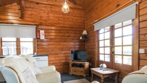 A wooden cabin interior with a sofa, a television, a small table, and a window. - Lodge 7 - Bron Eifion (Criccieth)