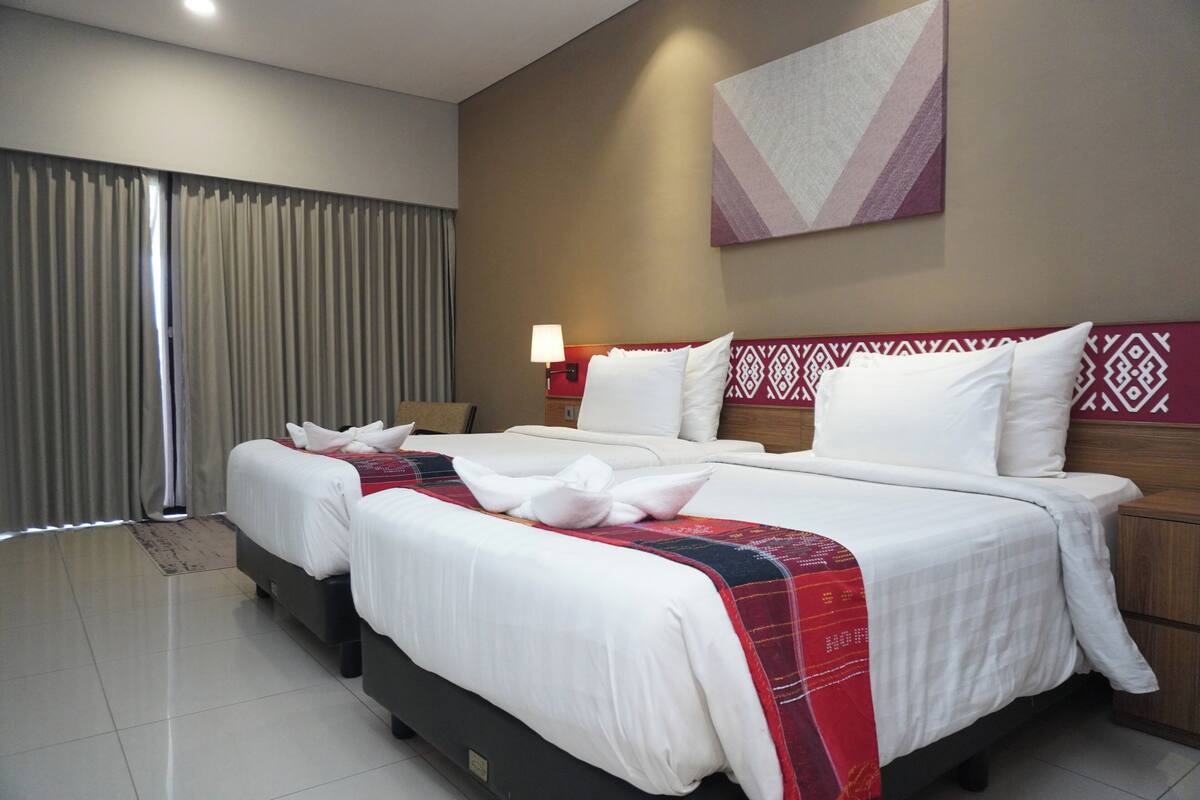A hotel room with two beds, a large window with curtains, a wall-mounted artwork, and a nightstand with a lamp.