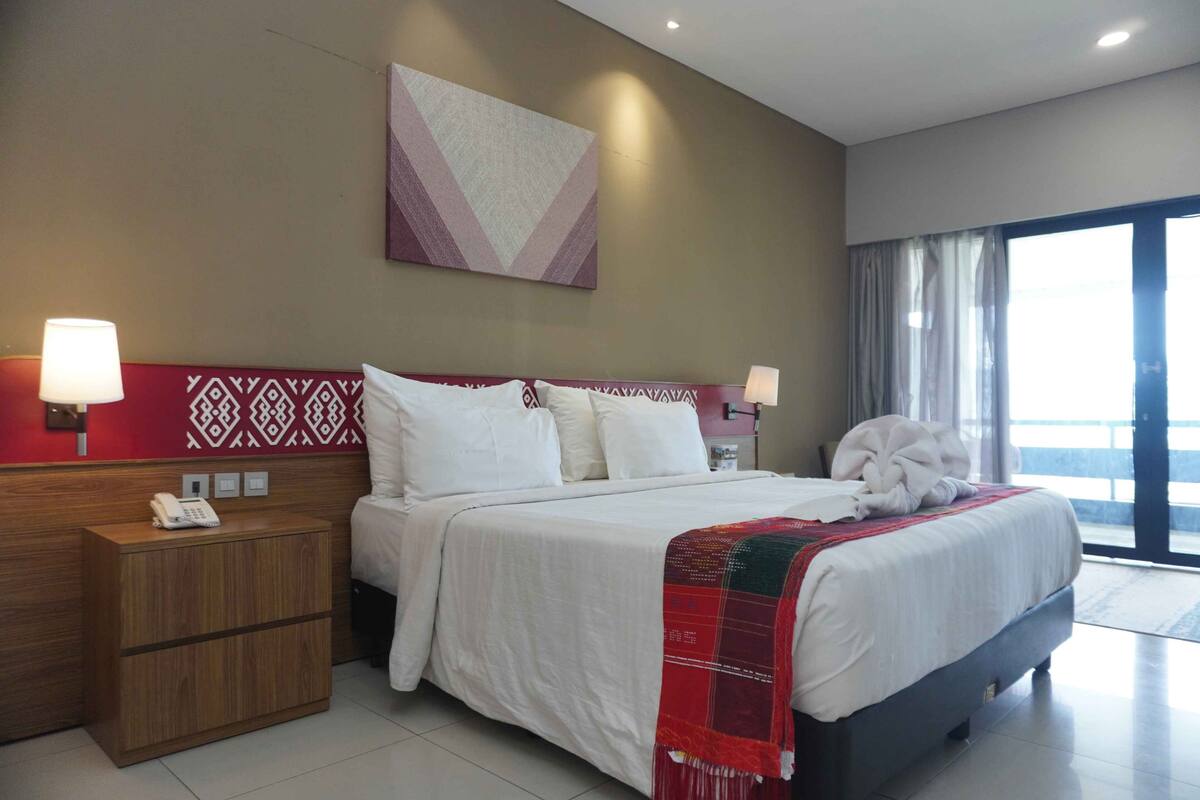 A hotel room with a large bed, bedside lamps, a wooden nightstand with a phone, a red and white patterned wall, and a large window with curtains.