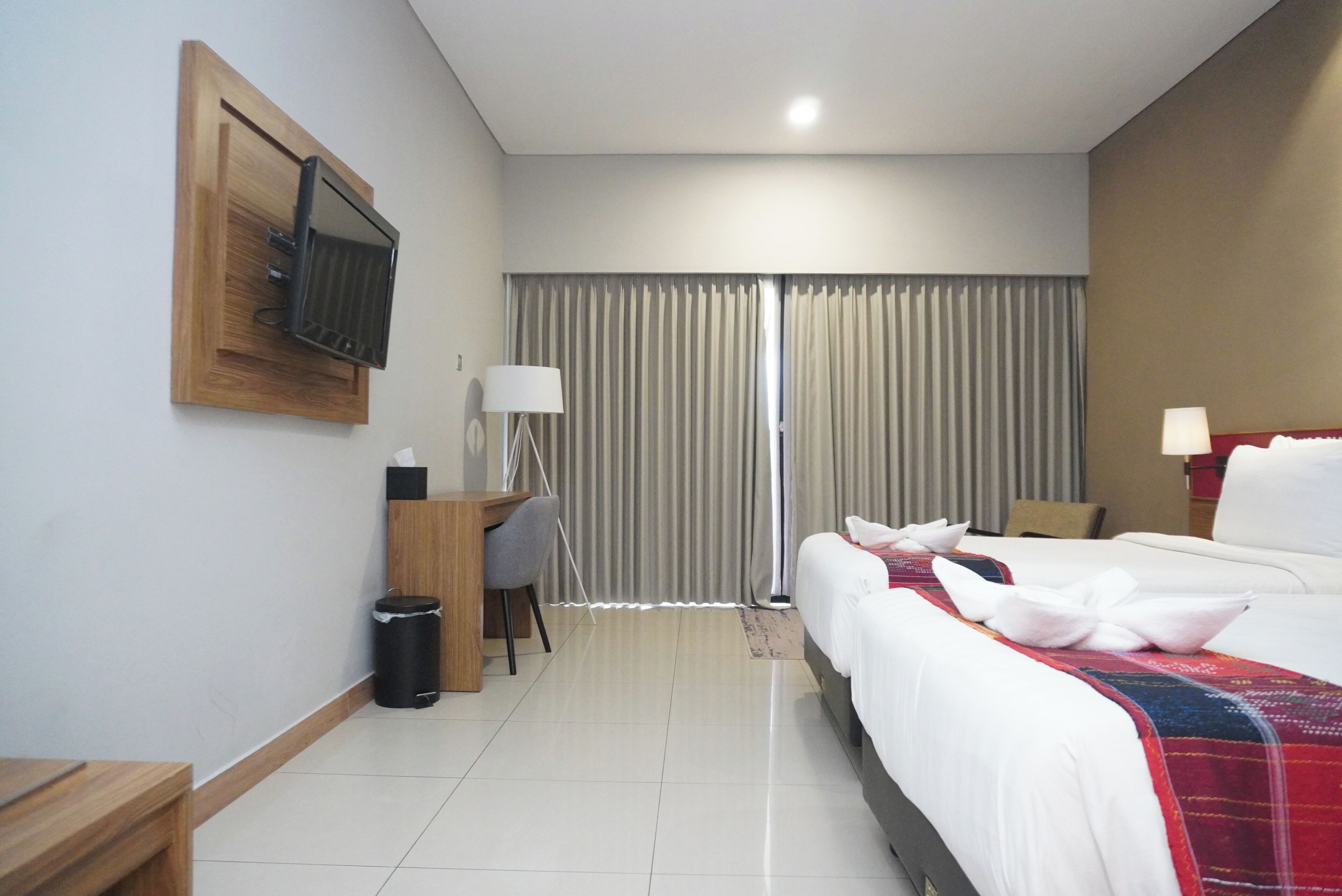 Habitación de hotel con cama, escritorio, silla, televisor montado en la pared y ventana con cortinas.