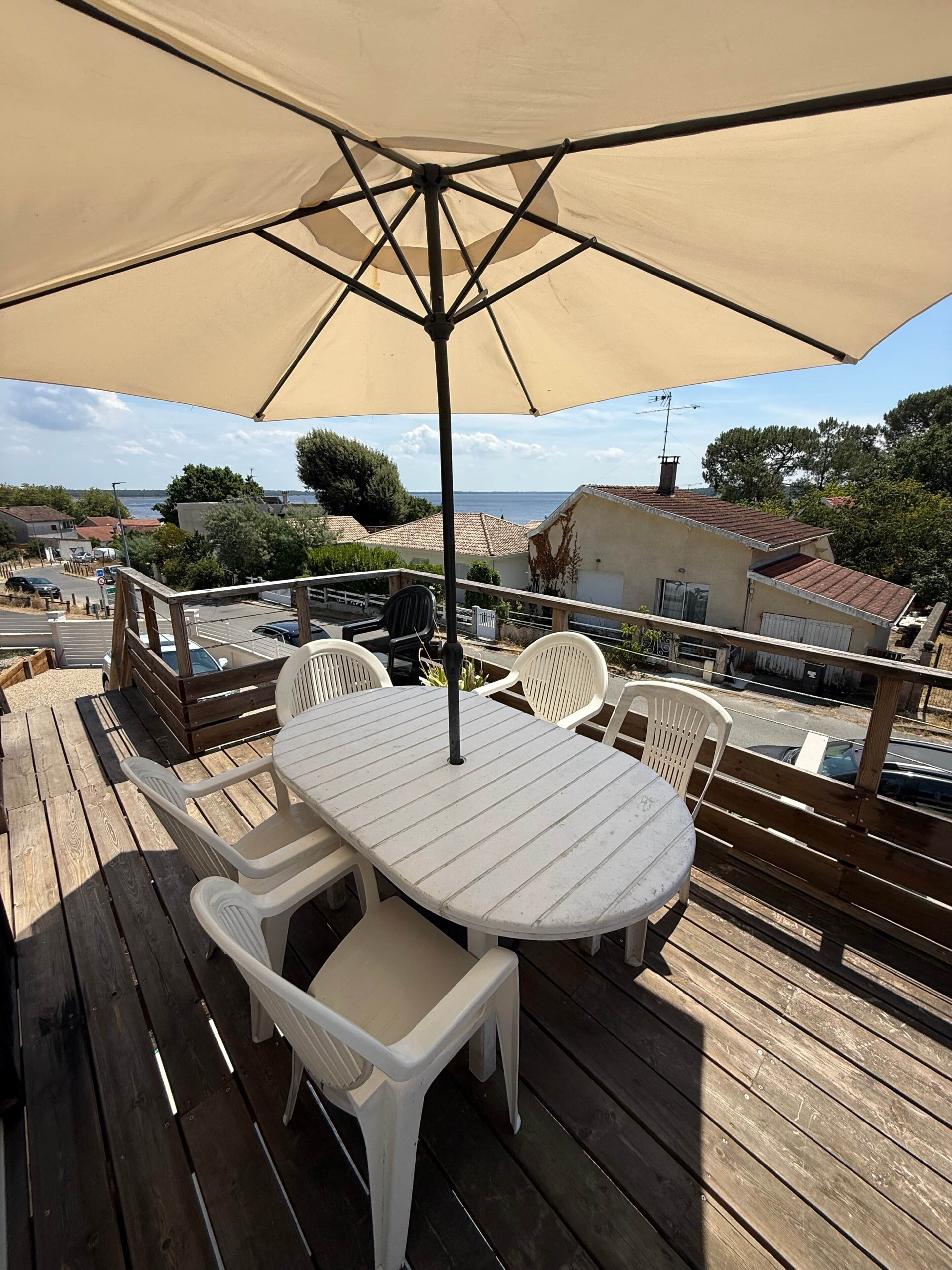A wooden deck with a table and chairs under a canopy, overlooking a coastal area with buildings and trees.