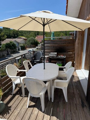 A wooden deck with a round table and white plastic chairs, an umbrella, and a grill. - House with panoramic lake view (Lacanau)