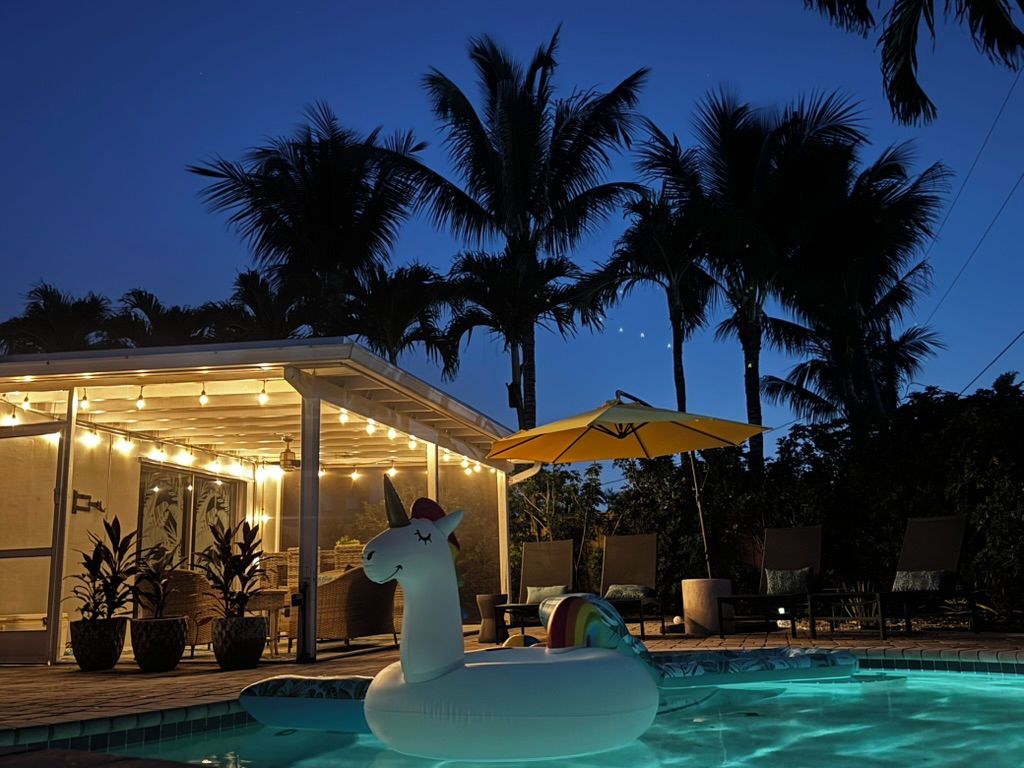 Ein Poolbereich mit einem Einhorn-Schwimmpfiffi, Palmen und einer überdachten Sitzgelegenheit mit Lichterkette.