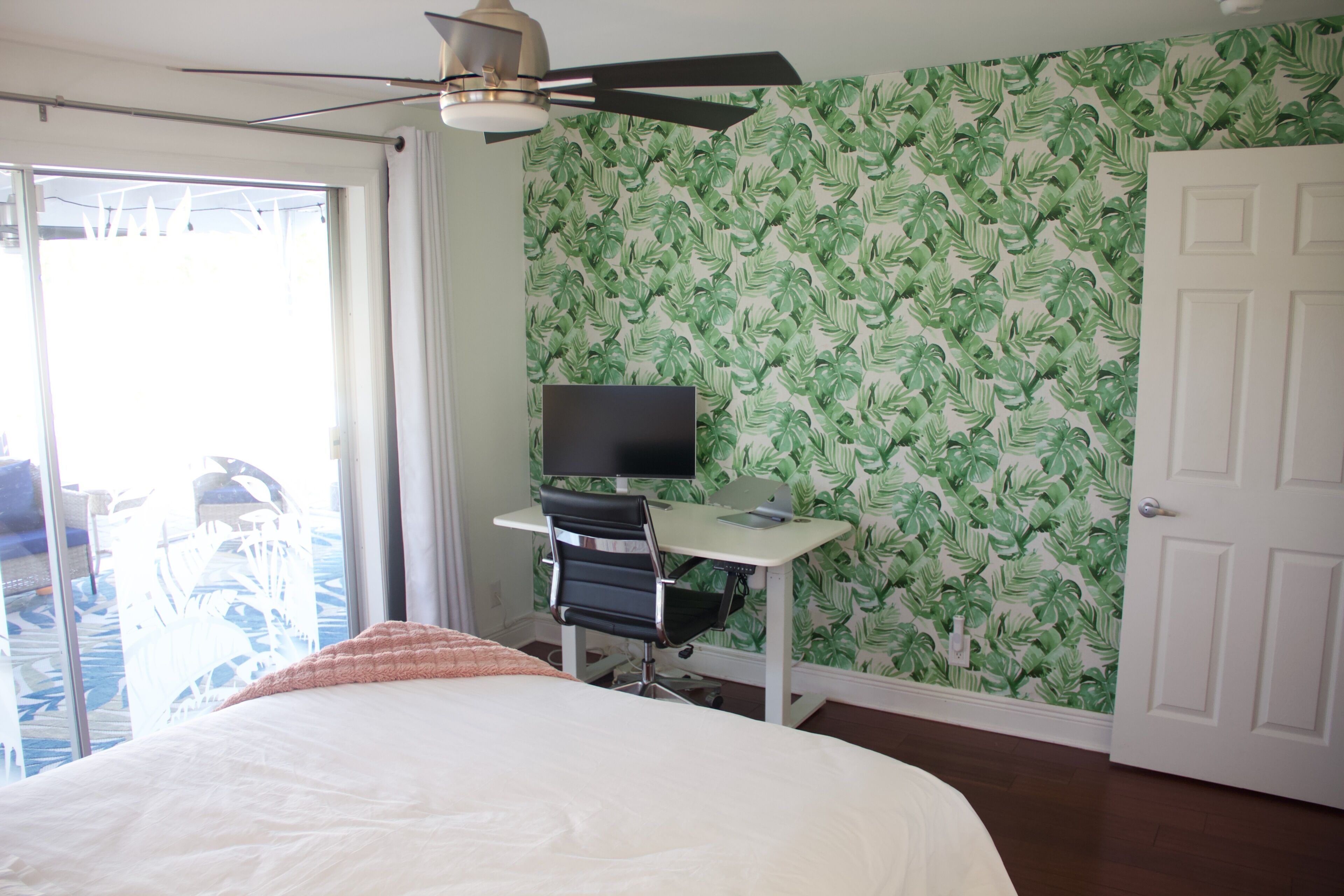 Une chambre à coucher avec un lit, un bureau avec une télévision, un ventilateur de plafond et une porte.