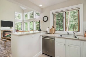 A kitchen with a fireplace, a clock, and a view of trees through large windows. - Modern Cabin, Ski-In/Out, Heated Pool Avail! (Boyne Falls)