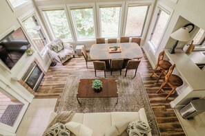 A spacious living area with a large window, a sofa, a coffee table, and a dining table with chairs. - Modern Cabin, Ski-In/Out, Heated Pool Avail! (Boyne Falls)