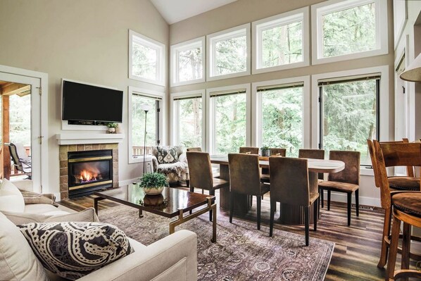 A living room with a fireplace, a television, and a dining area with chairs and a table. - Modern Cabin, Ski-In/Out, Heated Pool Avail! (Boyne Falls)
