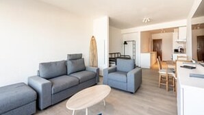 En moderne stue med en grå sofa, et rundt bord og et køkken i baggrunden.