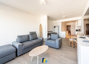 Ein modernes Wohnzimmer mit einem grauen Sofa, einem kleinen runden Tisch und einer Küchenzeile im Hintergrund.