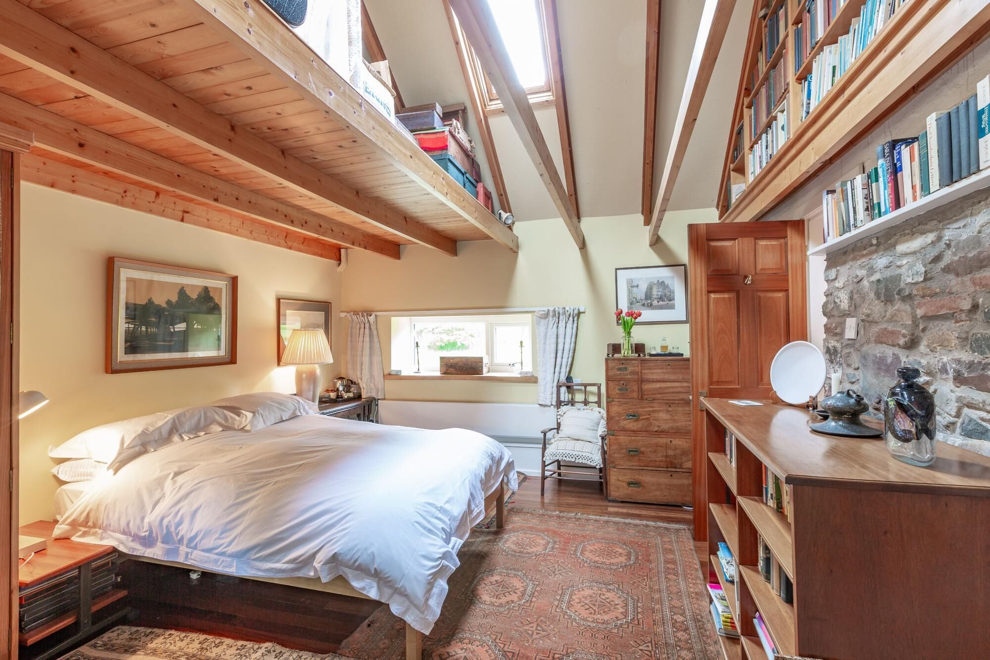 A bedroom with a loft bed, wooden beams, a stone wall, a desk, and a bookshelf.