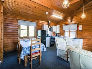A wooden cabin interior with a dining table, chairs, a sofa, a kitchen area, and a ceiling light fixture.
