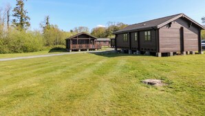 A wooden cabin with a porch, surrounded by a grassy field and trees. - Lodge 2 - Bron Eifion (Criccieth)
