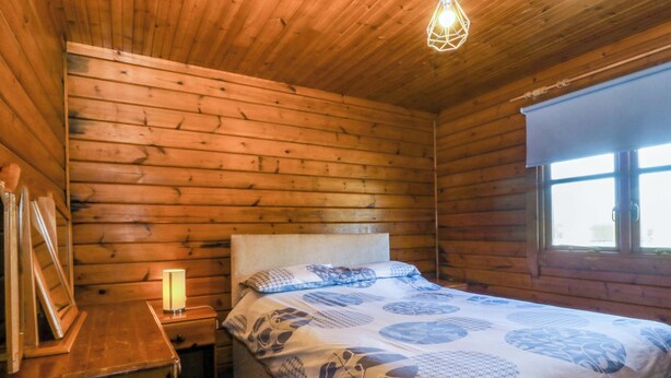 A wooden cabin bedroom with a bed, bedside table, lamp, and window. - Lodge 2 - Bron Eifion (Criccieth)