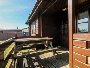 A wooden building with a covered patio area, a wooden bench, and a table. - Lodge 4 - Bron Eifion (Criccieth)
