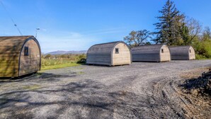 Three small, wooden, semi-circular structures with a flat roof, arranged in a row on a gravel surface, with a clear blue sky above and a tree to the right. - Bron Eifion Pod 2 (Criccieth)