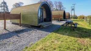 A row of wooden pod-style accommodations with a gravel ground, a picnic table, and a wooden fence. - Bron Eifion Pod 3 (Criccieth)