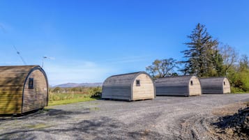 Three small, wooden, semi-circular structures with a flat roof, arranged in a row on a gravel surface, with a clear blue sky above and a tree to the right.