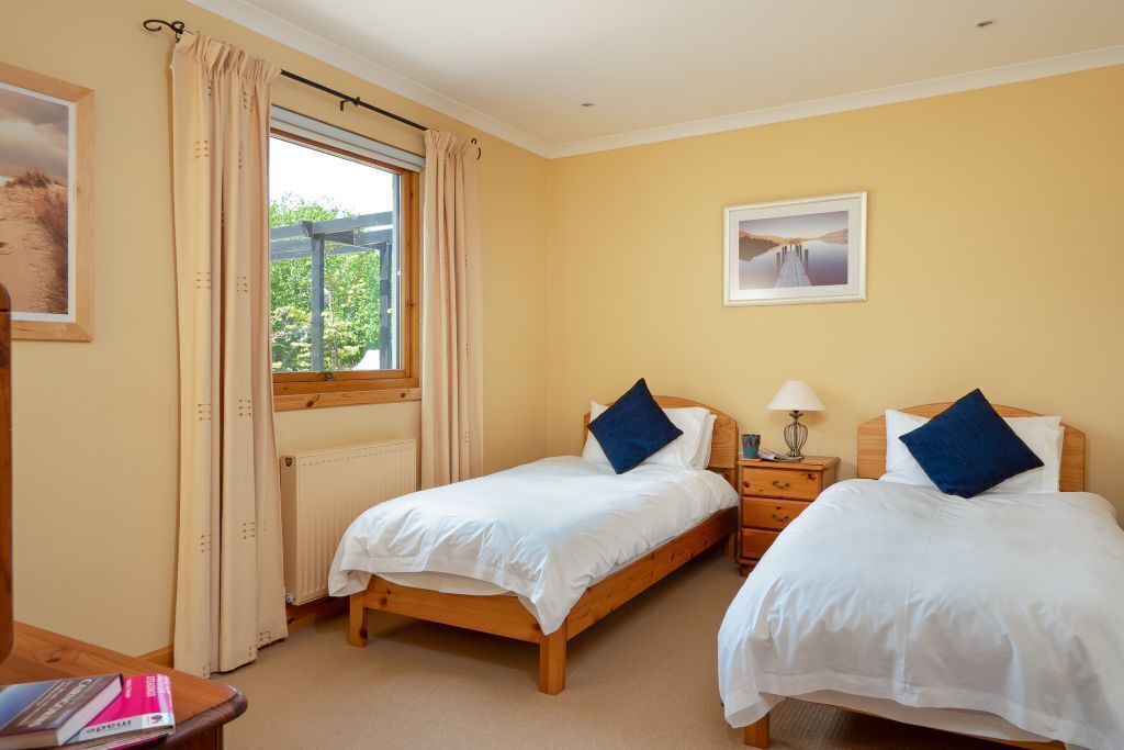 A room with two beds, a window with curtains, a radiator, a bedside table with a lamp, and a framed picture on the wall.