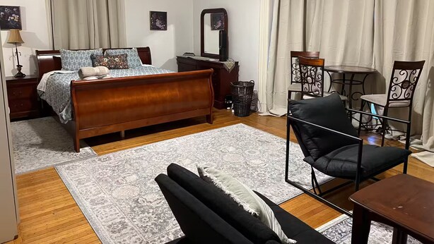 Ein Schlafzimmer mit einem hölzernen Bett, einem kleinen Tisch, einem Stuhl, einem Spiegel und einem Fenster mit Vorhängen.