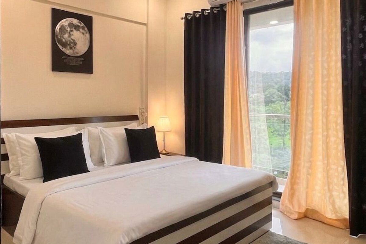 Habitación de hotel con cama, ventilador de techo, equipo de aire acondicionado, ventana con cortinas y un cuadro en la pared.