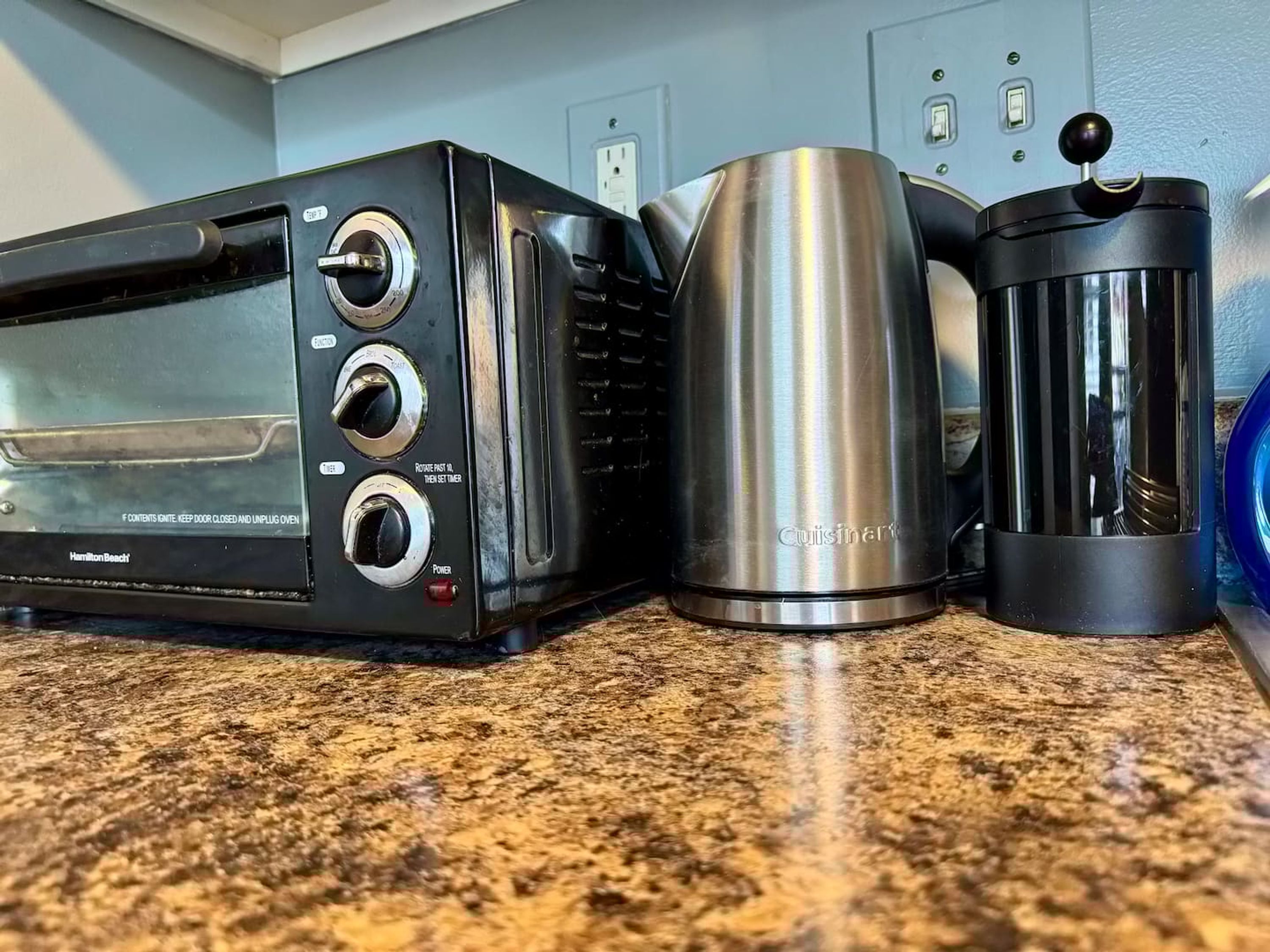 Una cocina con horno tostador, hervidor de acero inoxidable y cafetera sobre una encimera de granito.