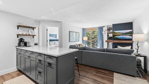 Eine moderne Küche mit Kochinsel, grauen Schränken und einem grauen Sofa im Wohnbereich mit Fernseher und Lampe.