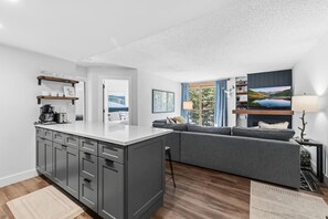 A modern kitchen with a central island, grey cabinets, and a grey sofa in a living area with a TV and lamp. - Breck Base Condo with Pool + Hot Tub (Breckenridge)