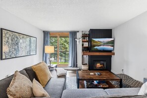 A living room with a sofa, coffee table, fireplace, TV, and artwork. - Breck Base Condo with Pool + Hot Tub (Breckenridge)
