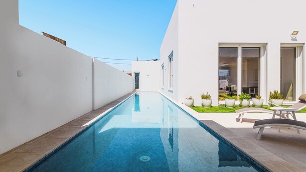 A narrow swimming pool with a clear blue water surface, surrounded by white walls and a small garden area with potted plants and a lounge chair. - Villa Jannah -Luxury Halal Retreat (Quinta do Anjo)