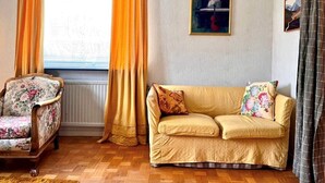 A room with a wooden floor, a floral armchair, a sofa with floral cushions, a radiator, and a window with curtains. - 7 Person Holiday Home in Virserum (Virserum)