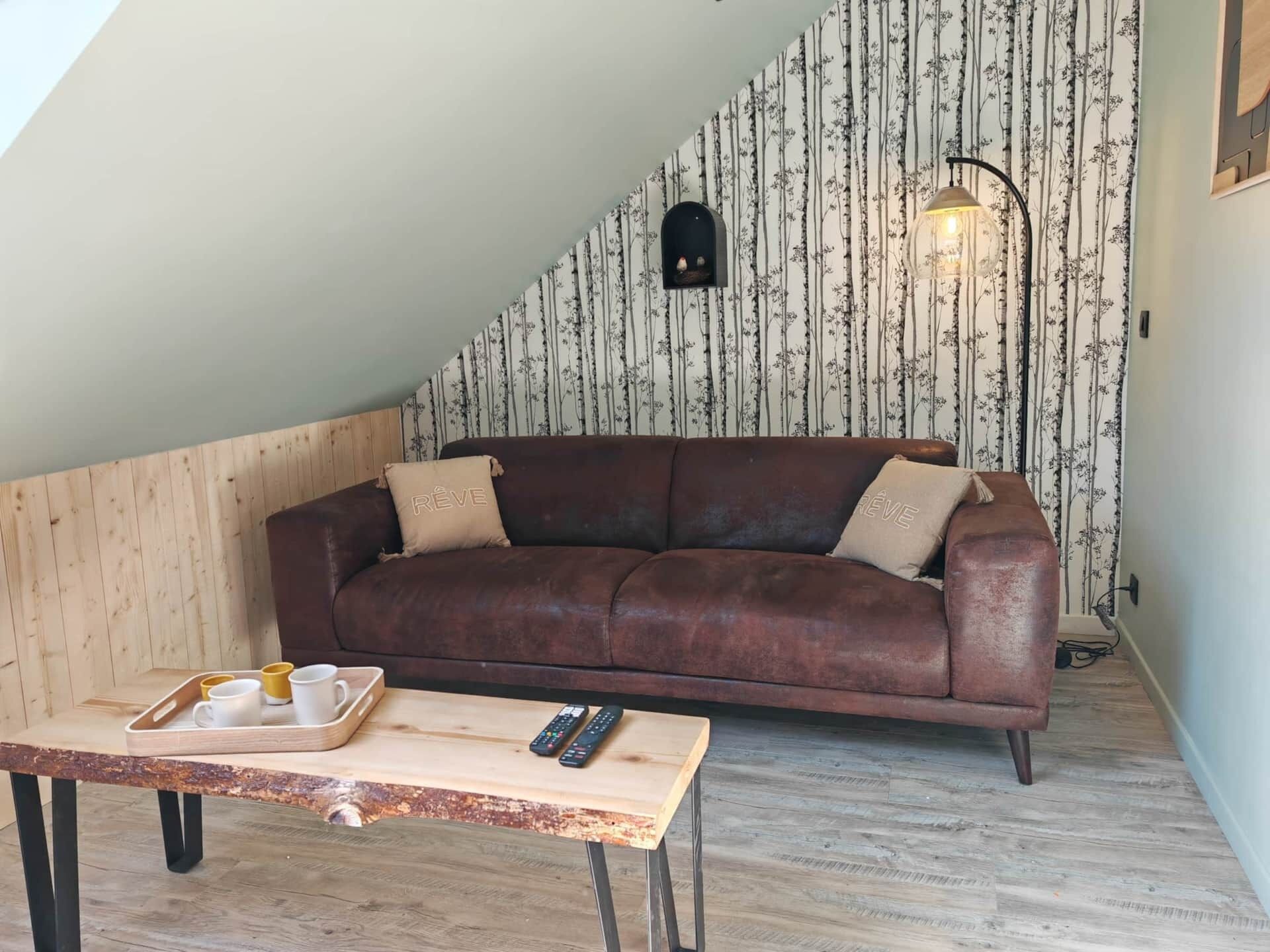A cozy loft bedroom with a brown leather sofa, a wooden table with a tray of mugs, and a sloped ceiling with a skylight.