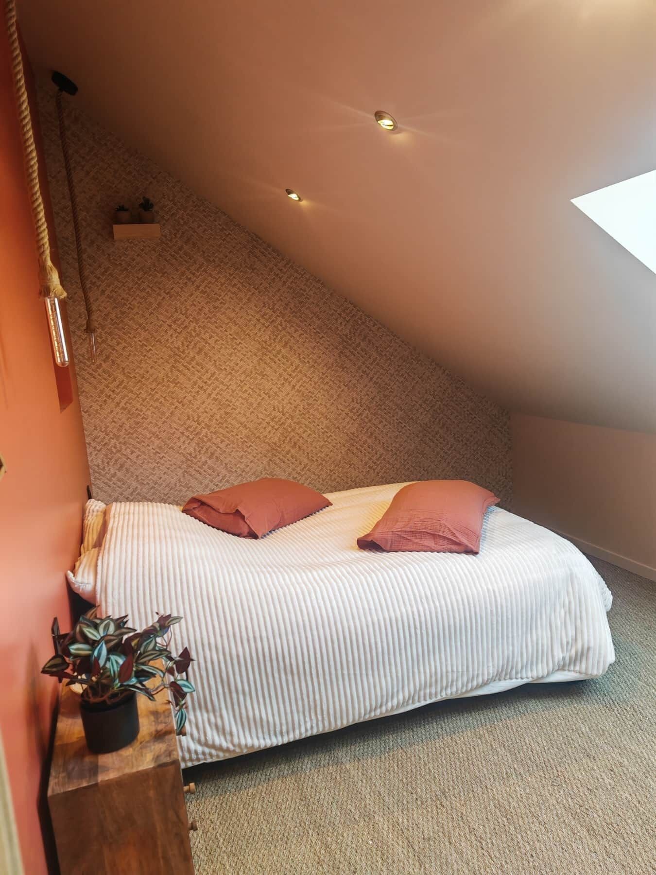 A bedroom with a sloped ceiling, a bed with white and pink bedding, a small plant on a wooden nightstand, and a ceiling light fixture.