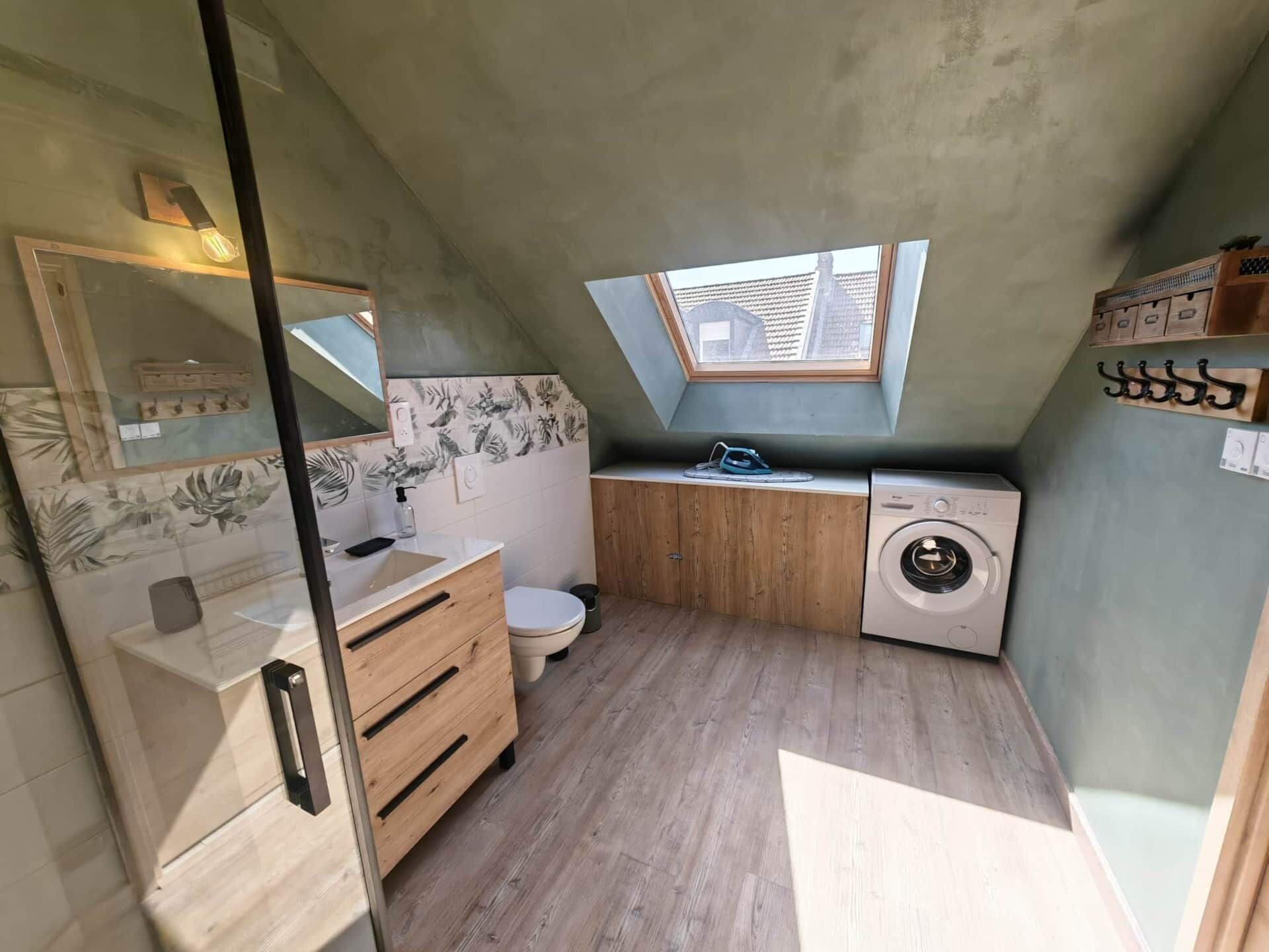 A compact, modern bathroom with a skylight, wooden cabinetry, a toilet, a washing machine, and a mirror.