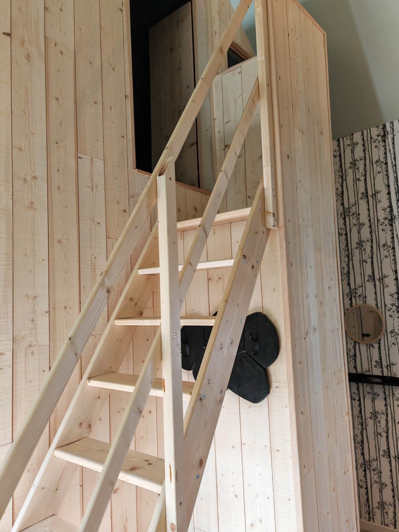 Un escalier en bois, avec une petite ouverture en haut, qui mène à une pièce dont les murs sont recouverts d’un papier peint à motifs.