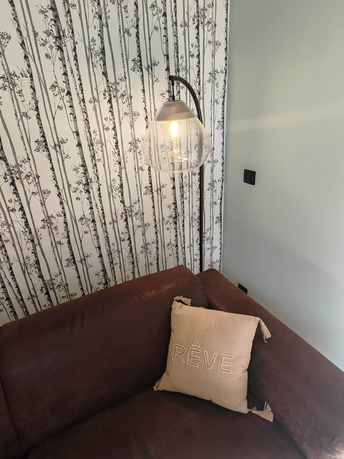 En brun soffa med en beige kudde med texten ”REVE” på, placerad framför en tapet med vertikala trädmönster.