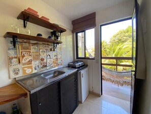 A kitchen with a sink, a small refrigerator, and a window with a view of greenery. - Kailua apartamentos e suítes  (Ipojuca)