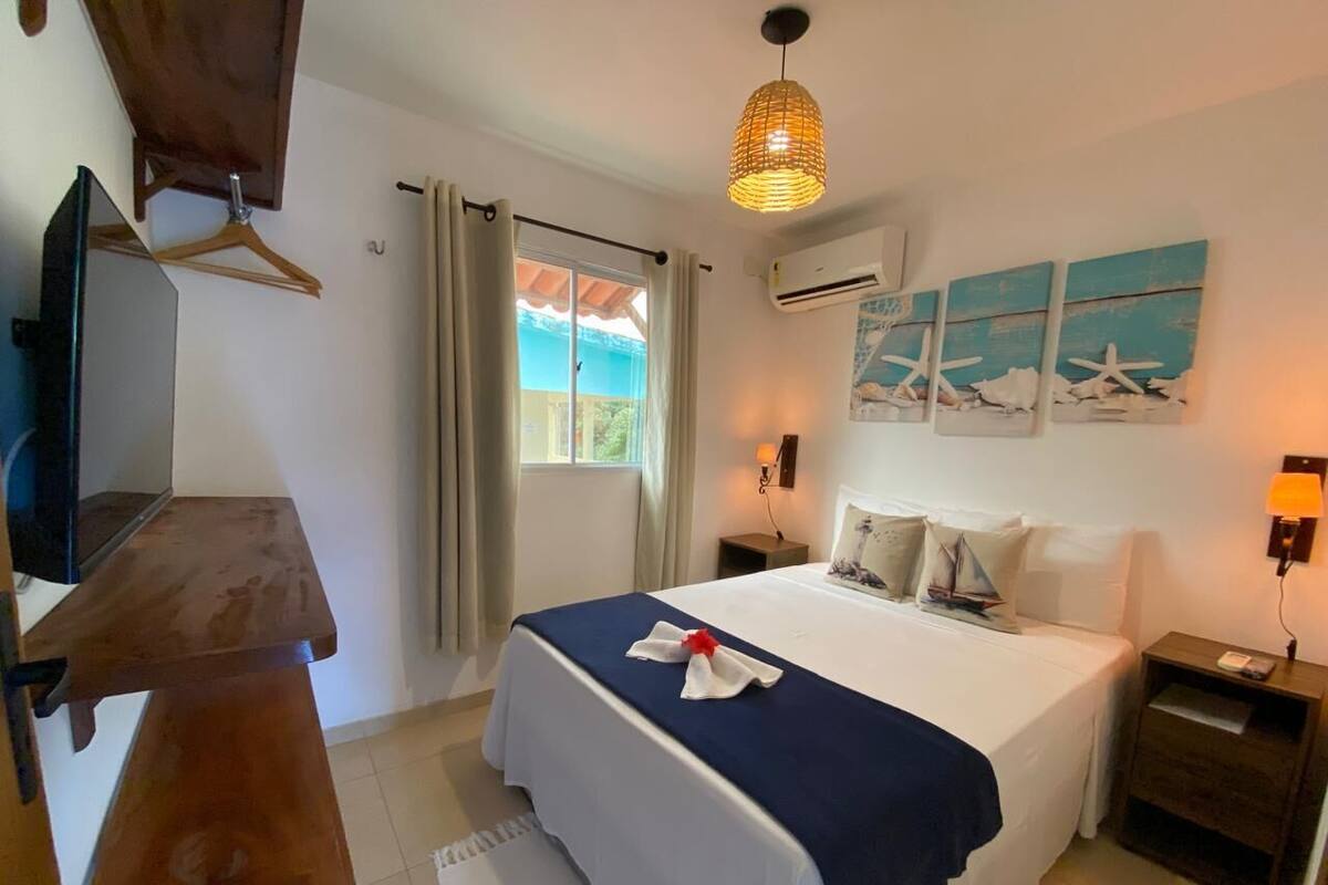 Une chambre à coucher comprenant un lit, des tables de chevet, un luminaire suspendu, une fenêtre avec des rideaux et des œuvres d’art murales.