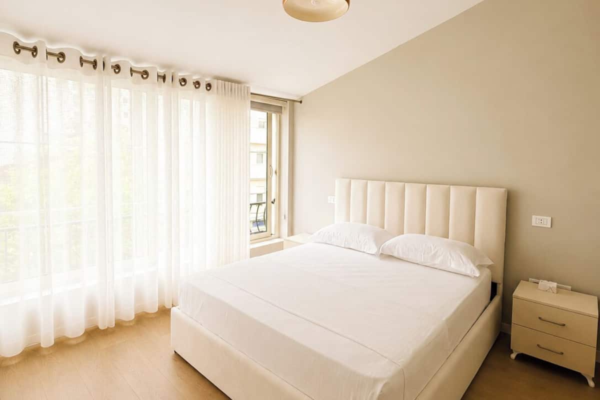 Quarto com uma cama grande, uma mesa de cabeceira e uma janela com cortinas leves.