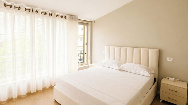 A bedroom with a large bed, a bedside table, a nightstand, and a window with sheer curtains. - Kein Hotel (Durres)