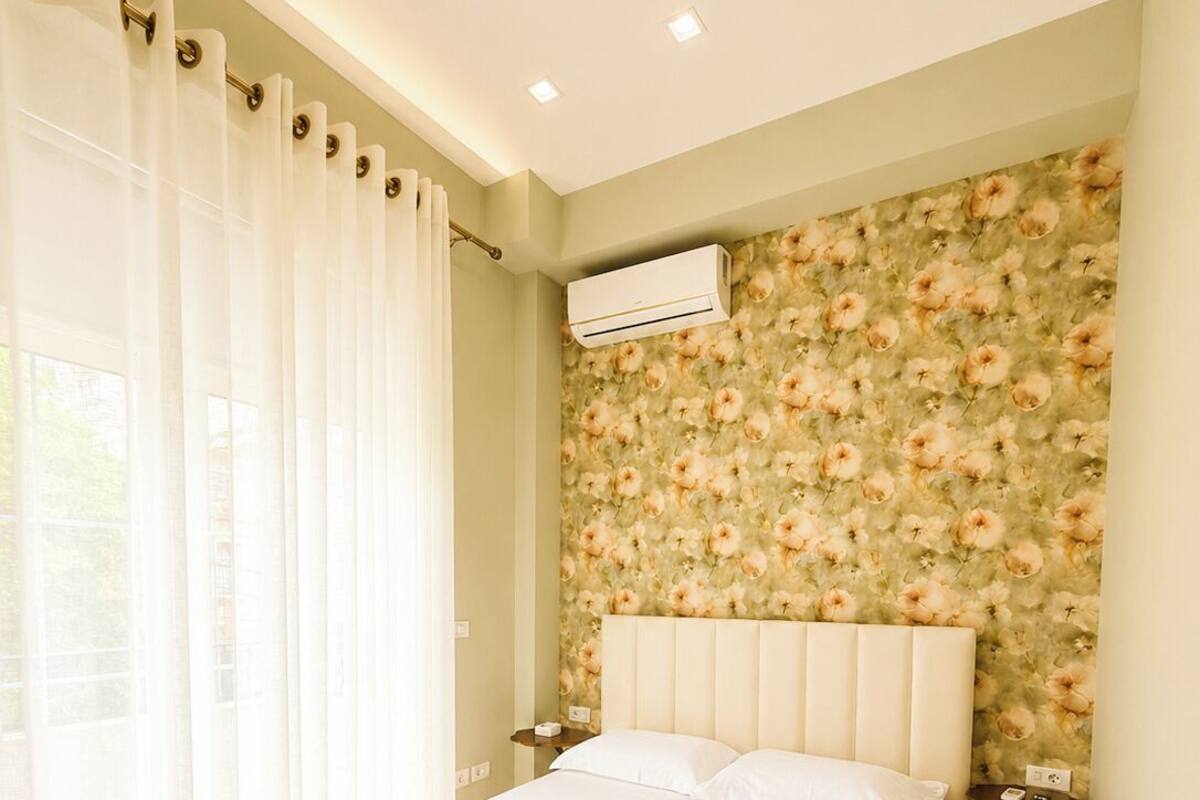 Quarto com cama, cortinas, papel de parede floral, aparelho de ar condicionado e mesa de cabeceira.