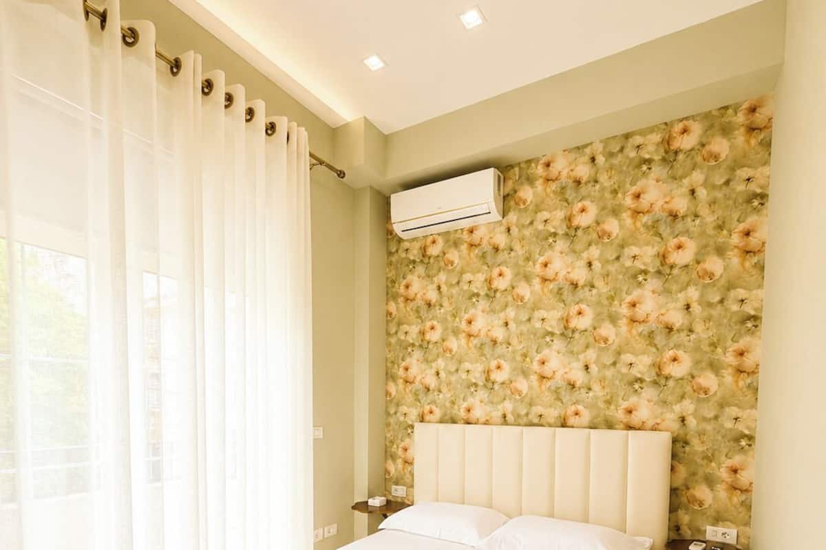 Quarto com cama, cortinas, papel de parede floral, aparelho de ar condicionado e mesa de cabeceira.