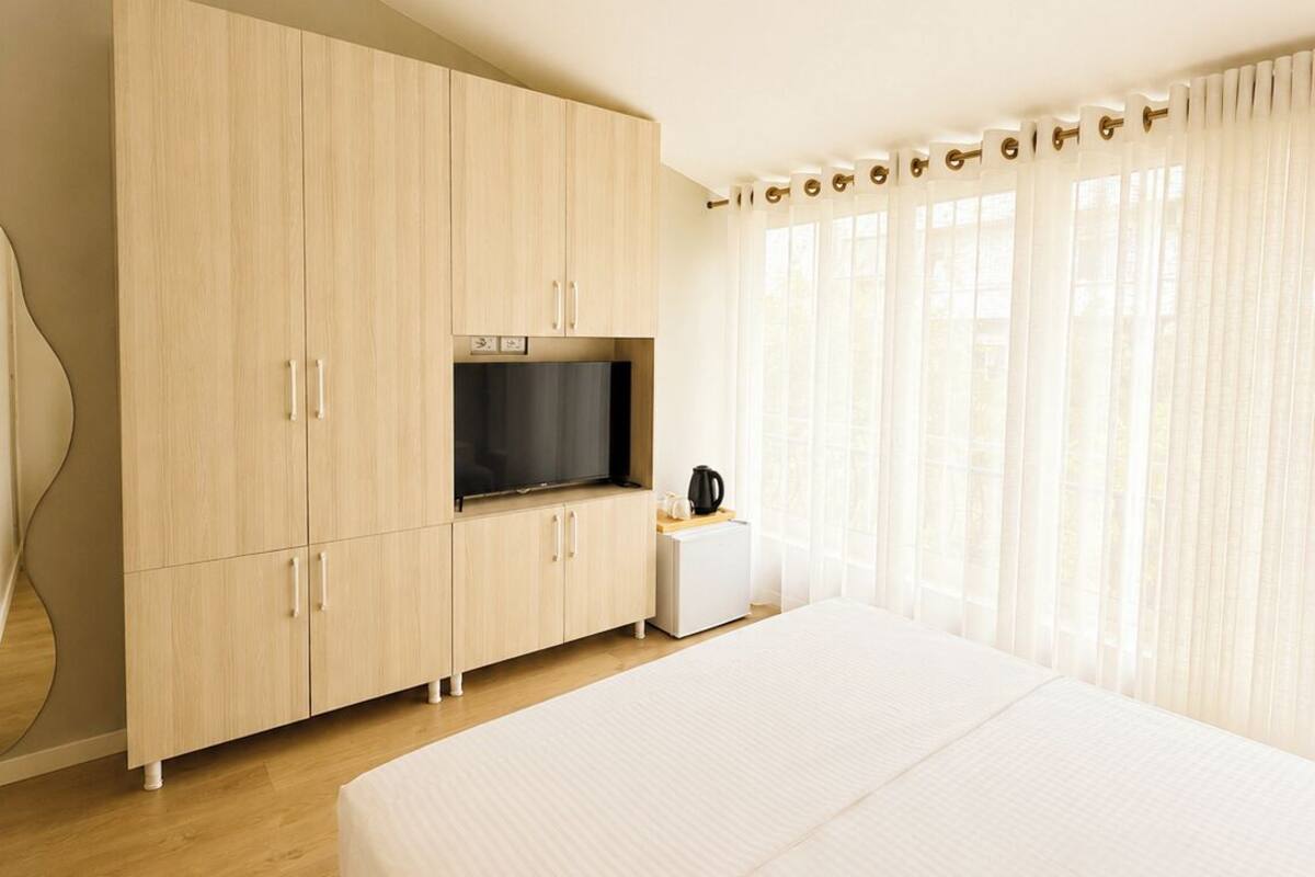 Quarto com uma cama grande, uma televisão em um armário de madeira, uma mesinha com uma chaleira e uma janela com cortinas.