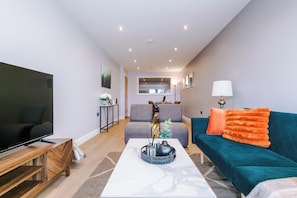 Ein modernes Wohnzimmer mit Flachbildfernseher, Couchtisch, einem taubenfarbenen Sofa mit orangefarbenen Kissen und einem Essbereich im Hintergrund.