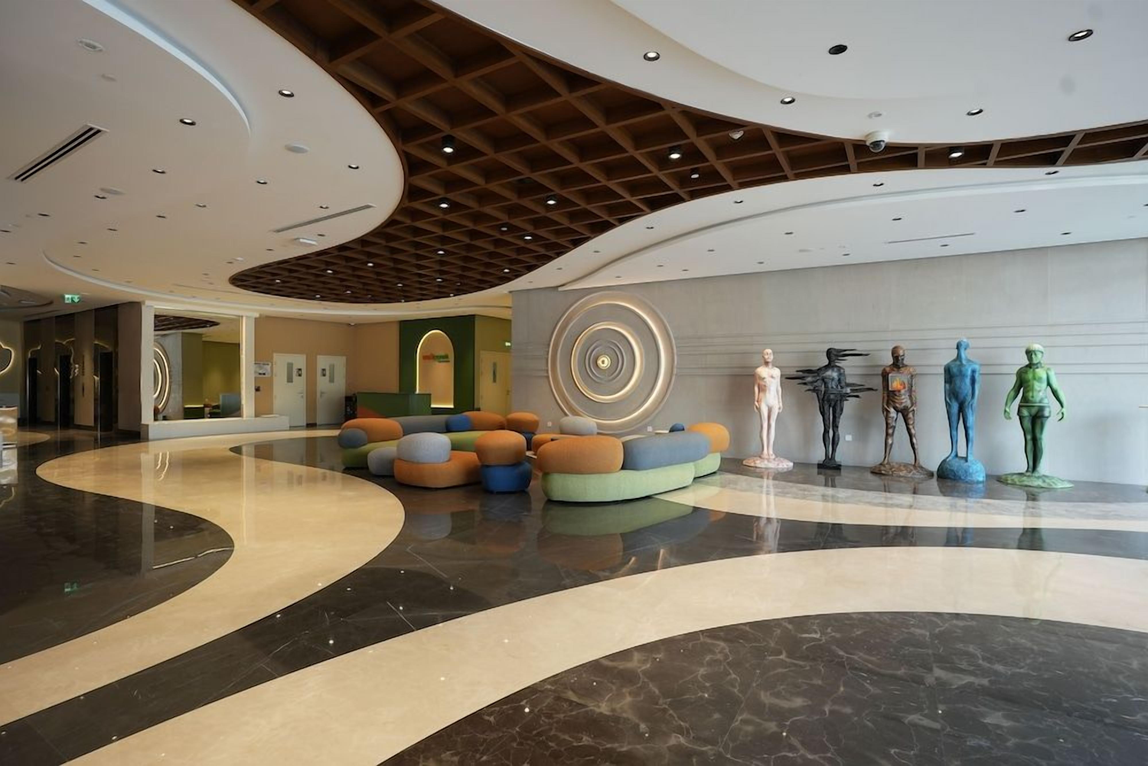 A modern lobby with a curved floor design, a central seating area with colorful cushions, and a row of statues on the wall.